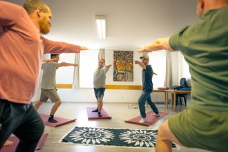 Ein Männer-Yogakurs in einem hellen Raum. Die Teilnehmer stehen auf bunten Matten und führen eine Armübung aus. Im Hintergrund ist ein farbenfrohes Gemälde zu sehen.