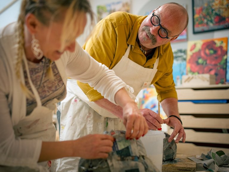 Ein Mann mit gelbem Hemd und Brille hilft einer Frau beim Basteln in einem Kunststudio. Beide tragen weiße Schürzen. Sie arbeiten gemeinsam an einem Objekt auf dem Tisch, während im Hintergrund bunte Gemälde hängen.