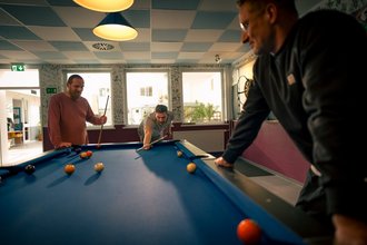 Drei Männer spielen eine Partie Poolbillard. Ein Mann beugt sich über den blauen Tisch, um einen Stoß auszuführen, während die anderen beiden ihm aufmerksam zuschauen.