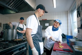 Eine ältere Köchin schneidet Kartoffeln auf einem roten Brett, während ein junger Mann daneben steht und ihr aufmerksam zuschaut. Im Hintergrund rührt ein weiterer Koch in einem großen Topf auf dem Herd.