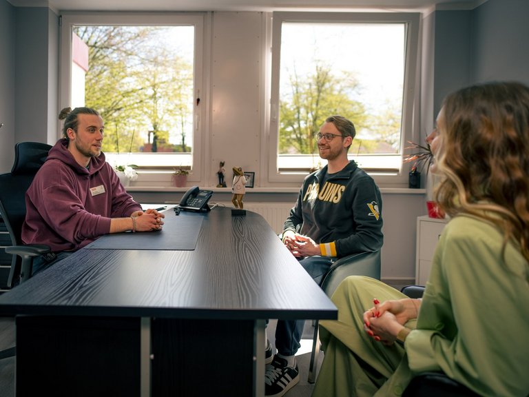 Drei Personen unterhalten sich an einem dunklen Schreibtisch in einem hellen Büro. Links sitzt ein Mann im burgunderroten Hoodie, mittig ein Mann im schwarzen Hoodie und rechts eine Frau in hellgrüner Kleidung. Im Hintergrund sind Fenster mit Blick auf Bäume zu sehen.