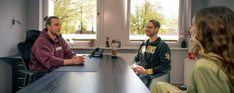 Drei Personen unterhalten sich an einem dunklen Schreibtisch in einem hellen Büro. Links sitzt ein Mann im burgunderroten Hoodie, mittig ein Mann im schwarzen Hoodie und rechts eine Frau in hellgrüner Kleidung. Im Hintergrund sind Fenster mit Blick auf Bäume zu sehen.