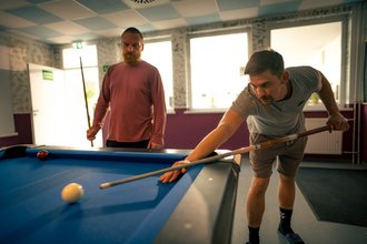 Zwei Männer spielen Billard. Ein Mann im grauen T-Shirt beugt sich konzentriert über den blauen Tisch und zielt mit dem Queue. Im Hintergrund steht ein bärtiger Mann und beobachtet das Spiel.