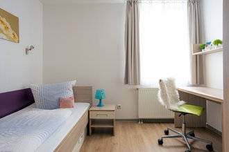 Ein helles Einzelzimmer mit hellem Holzboden. Links steht ein Bett mit blauer Bettwäsche und ein Nachttisch mit einer blauen Lampe. Rechts befindet sich ein Schreibtisch unter einem Regal mit kleinen Pflanzen. Davor steht ein grüner Bürostuhl mit weißem Fellüberwurf. Ein großes Fenster mit Vorhängen sorgt für viel Tageslicht.