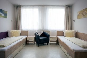 Ein helles Schlafzimmer mit zwei Einzelbetten, die mit gelber Bettwäsche bezogen sind. In der Mitte steht ein dunkelblauer Sessel zwischen zwei Nachttischen. Im Hintergrund befindet sich ein großes Fenster mit weißen Vorhängen.