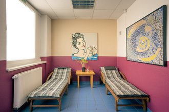 Ein Ruheraum mit zwei Holzliegestühlen auf blauem Fliesenboden. Dazwischen steht ein kleiner Holztisch mit einer Pflanze. An den Wänden hängen ein Pop-Art-Gemälde einer Frau und ein großes abstraktes Mosaikbild.