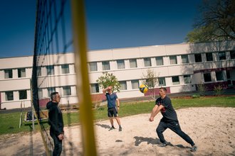 Drei Männer spielen Beachvolleyball auf einem Sandplatz. Ein Spieler im rechten Vordergrund bereitet sich vor, den gelb-blauen Ball zu empfangen, während zwei weitere Männer das Spiel beobachten. Im Hintergrund steht ein weißes Gebäude.