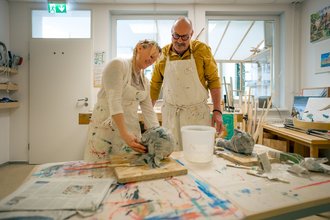 In einem hellen Atelier arbeiten ein Mann und eine Frau gemeinsam an einer Skulptur aus Papiermaché. Beide tragen schmutzige weiße Schürzen. Die Frau formt das Objekt, während der Mann sie beobachtet. Der Tisch ist mit Farben und Werkzeug bedeckt.
