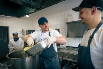 In einer Großküche füllt ein Koch Kartoffelwürfel aus einer Metallschale in einen großen Topf. Ein Kollege rechts im Bild beobachtet ihn lächelnd. Im Hintergrund ist eine weitere Mitarbeiterin in Kochkleidung zu sehen.