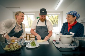 Drei Personen stehen in einer Küche und bereiten gemeinsam Obst zu. Ein Mann mit Schürze schneidet eine Banane, während eine Frau links und eine ältere Dame rechts lächelnd dabei zuschauen. Auf der Arbeitsplatte stehen Schüsseln mit geschnittenem Obst und Trauben.