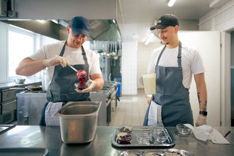 Zwei Köche in einer professionellen Küche bereiten Desserts zu. Einer füllt rote Fruchtsoße in Gläser, während der andere eine Kanne mit Sahne hält und lächelnd zuschaut.