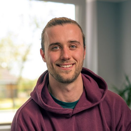Ein junger Mann mit kurzem Haar und Bartstoppeln sitzt lächelnd in einem hellen Büro. Er trägt einen burgunderroten Hoodie und blickt freundlich direkt in die Kamera. Im Hintergrund sind ein Fenster und ein Schreibtisch unscharf zu erkennen.