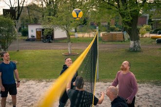 Fünf Männer spielen Volleyball auf einem Sandplatz im Freien. Ein gelb-blauer Ball schwebt über dem Netz, während ein Spieler ihn schlägt. Die anderen Männer beobachten das Spiel aufmerksam.