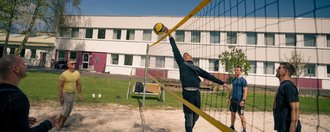 Männer spielen Beachvolleyball auf einem Sandplatz an einem sonnigen Tag. Ein Spieler springt hoch, um den Ball über das Netz zu schlagen, während andere Mitspieler das Spiel beobachten. Im Hintergrund ist ein weißes Gebäude zu sehen.