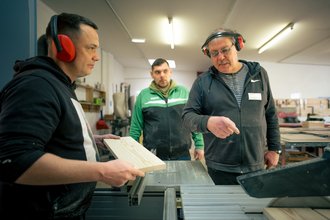 Drei Männer in einer Holzwerkstatt. Ein Ausbilder mit Gehörschutz erklärt einem jungen Mann die Handhabung einer Kreissäge, während dieser ein Holzbrett positioniert. Ein weiterer Mann beobachtet die Situation im Hintergrund.