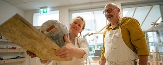 Zwei Künstler in schmutzigen weißen Kitteln stehen in einer Werkstatt. Die Frau hält ein großes Kunstobjekt aus Holz und Papiermache hoch und lächelt, während der Mann mit Brille und Schnurrbart ihr zufrieden zuschaut.