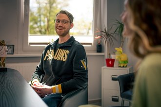 Ein junger Mann mit Brille und Bart sitzt lächelnd in einem Büro. Er trägt einen schwarzen Kapuzenpullover mit der gelben Aufschrift „Penguins“. Er blickt freundlich auf eine Frau im Vordergrund, die unscharf zu sehen ist, was auf ein Gespräch hindeutet.
