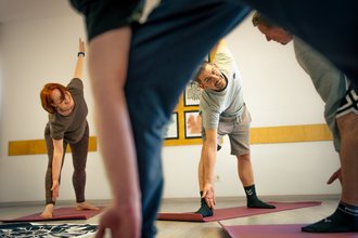 Ein Mann und eine Frau mit roten Haaren lächeln fröhlich, während sie auf Yogamatten eine seitliche Dehnübung ausführen. Im Vordergrund sind die unscharfen Beine anderer Teilnehmer zu sehen. Die Gruppe befindet sich in einem hellen Raum.