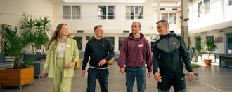 Vier Personen, eine Frau und drei Männer, gehen nebeneinander durch einen hellen Innenraum. Sie tragen Namensschilder und unterhalten sich. Die Frau trägt einen hellgrünen Anzug, die Männer legere Freizeitkleidung wie Hoodies und Trainingshosen.
