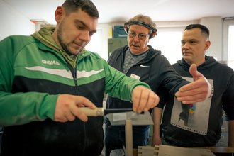 Drei Männer in einer Werkstatt. Ein junger Mann in grüner Jacke sägt Holz, während ein älterer Mann mit Gehörschutz ihn anleitet. Ein dritter Mann beobachtet aufmerksam. Die Szene zeigt eine praktische Unterweisung oder Ausbildung im Handwerk.