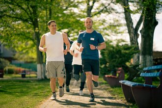 Zwei Männer joggen auf einem gepflasterten Weg in einem Park, gefolgt von zwei weiteren Läufern. Der Mann links trägt ein weißes ärmelloses Shirt, der Mann rechts ein blaues T-Shirt. Sie laufen an grünen Bäumen und Bänken vorbei.