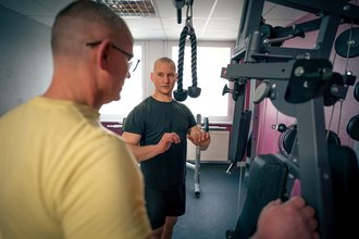 Ein Fitness-Trainer erklärt einem Kunden die Handhabung einer Trainingsmaschine. Der Trainer im schwarzen Shirt steht dem Kunden im gelben Shirt gegenüber und gestikuliert, um die richtige Übungsausführung zu demonstrieren.