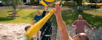 Männer spielen Beachvolleyball auf einem Sandplatz. Ein Spieler im gelben Shirt schlägt einen gelb-blauen Ball über das Netz, während ein Gegenspieler im schwarzen Oberteil versucht zu blocken. Weitere Spieler beobachten das Spiel bei sonnigem Wetter im Freien.