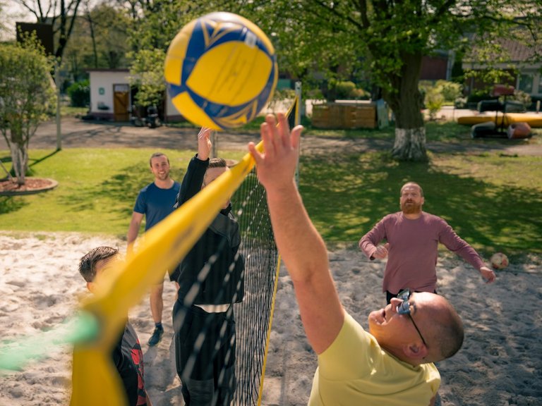 Männer spielen Beachvolleyball auf einem Sandplatz. Ein Spieler im gelben Shirt schlägt einen gelb-blauen Ball über das Netz, während ein Gegenspieler im schwarzen Oberteil versucht zu blocken. Weitere Spieler beobachten das Spiel bei sonnigem Wetter im Freien.