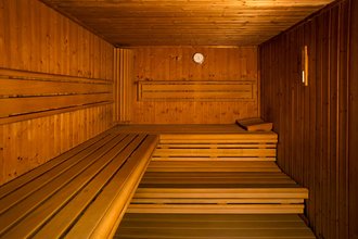 Das Bild zeigt den Innenraum einer leeren Holzsauna. Die Wände, Decke und gestaffelten Bänke bestehen aus hellem Holz. Warmes Licht beleuchtet den Raum. Auf der obersten Sitzbank liegt ein kleines Holzkissen. An der hinteren Wand hängt ein rundes Thermometer.