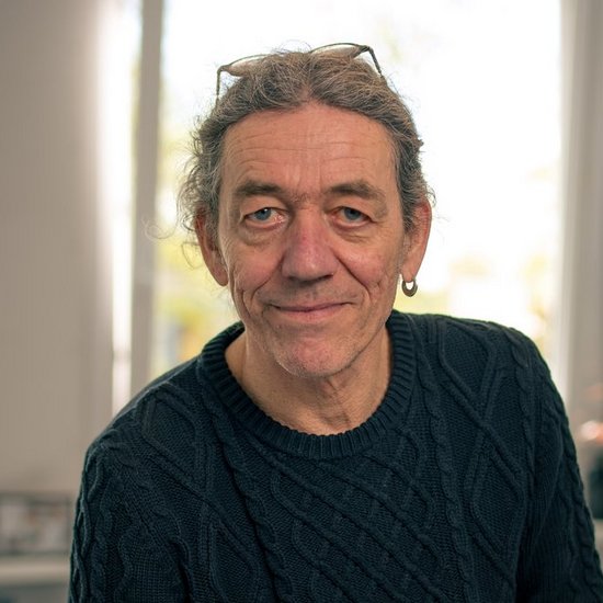 Ein lächelnder Mann mittleren Alters mit einer Brille auf dem Kopf und einem dunklen Strickpullover sitzt auf einem Bürotisch. Im Hintergrund sind ein helles Fenster und Pflanzen unscharf zu erkennen.
